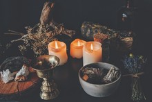 Messy Wiccan Witch Altar Working Space With Ingredients For A Spell. Dried Plants Nature Elements Branches And Lavender On A Table With Burning Candles, Sage Smudge Sticks, Gold Brass Incense Holder