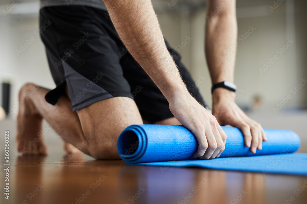Close-up of unrecognizable man standing on knees and rolling out exercise mat after yoga training in gym  - obrazy, fototapety, plakaty 