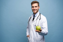 Portrait Of Young Male Doctor Holding Green Apple. Isolated On Light Blue.