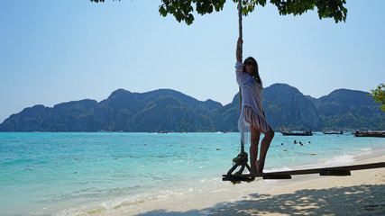 Beautiful girl in a white Cape and a black bathing suit on a swing. Swing on the beach. View of the island, beach with sand, blue water and green leaves of trees. Visible hills of the island.