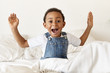 Emotional cute little African American boy sitting on white bed, screaming with mouth wide opened and gesturing actively, being in panic because he overslept, late for school. Human emotions