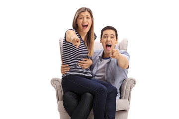 Canvas Print - Happy young couple in an armchair pointing at the camera and laughing