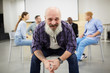 © Seventyfour - Portrait of bearded senior man smiling at camera while sitting in chair in front of therapy group, copy space
