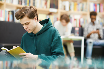 Wall Mural - Serious concentrated student guy in hoodie sitting in modern book store and reading book with interest