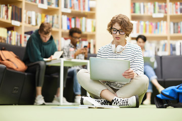 Wall Mural - Serious pensive curly-haired student girl with headphones on neck sitting with crossed legs on floor in library and analyzing online resources on laptop