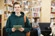 © Seventyfour - Cheerful confident handsome high school student with satchel standing in modern library and using digital tablet while looking at camera