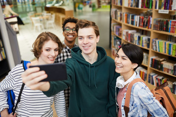 Wall Mural - Group of cheerful excited young multiethnic students in casual clothing standing in book store and taking selfie together on smartphone