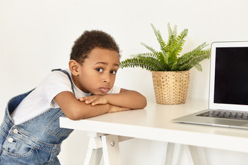 Wall Mural - Sad upset Afro American preschooler placing elbows on white table with open laptop computer with blank copyspace screen, looking at camera, having mournful expression, forbidden to watch cartoons