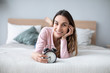 © BestForYou - Smiling young woman holding alarm clock lying on the bed while looking at camera.