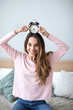 © BestForYou - Smiling young woman holding alarm clock sitting on the bed while looking at camera.