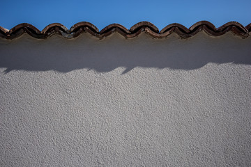 White plaster on the wall, background or texture, spring day, Pazardzhik region, Bulgaria. Top of the wall covered with two layers of ceramic roof tiles, Sun casting shadows on the surface