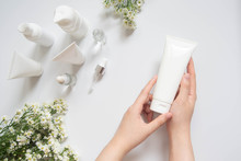 Young Female Hand Holding Blank White Squeeze Bottle Plastic Tube W/ Organic Natural Skincare Products And Flower On White Table. Packaging Of Lotion, Cream Or Serum. Beauty Cosmetic Skincare Concept.