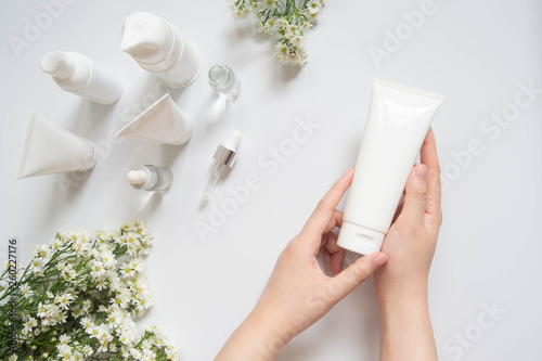Fotomural Young female hand holding blank white squeeze bottle plastic tube w/ organic natural skincare products and flower on white table