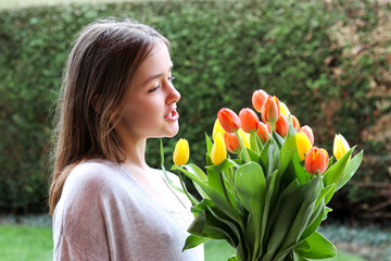 Beautiful smiling happy tween girl holding big bouquet of bright yellow and orange tulips talking to them outdoors in garden at warm spring day. Spring flowers