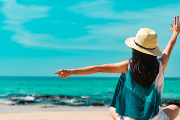 Wall Mural - Happy young woman wear straw hat sit and raised hand at sand beach. Relaxing and enjoy holiday at tropical paradise beach with green water. Girl in summer vacation. Summer vibes. Carefree concept.