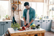 © gstockstudio - Homemade dinner. Handsome young man in casual wear cutting vegetables while standing in the kitchen at home