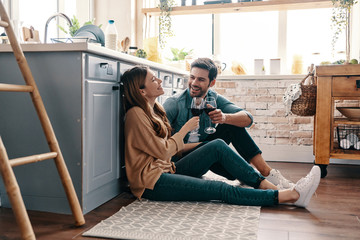 Sticker - Love is in the air. Beautiful young couple drinking wine while sitting on the kitchen floor at home