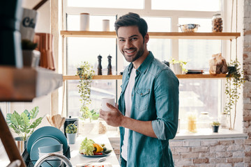 Morning filled with joy. Handsome young man in casual wear drinking coffee and smiling while standing in the kitchen at home