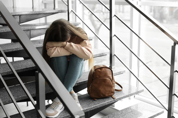 Wall Mural - Upset teenage girl with backpack sitting on stairs indoors. Space for text