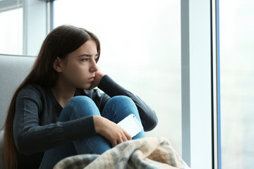 upset teenage girl with smartphone sitting at window indoors. space for text