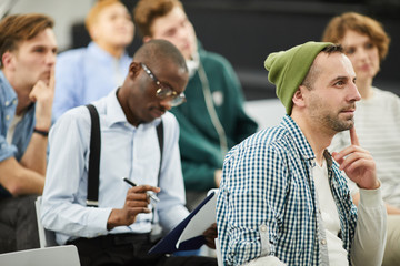 Group of pensive modern multiethnic students in casual clothing sitting in auditorium and listening to speaker at conference