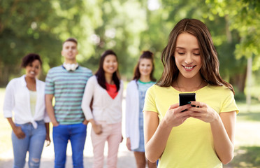 Wall Mural - technology and people concept - smiling young woman or teenage girl in blank yellow t-shirt using smartphone over group of friends summer park background