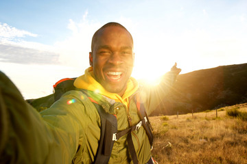 Wall Mural - Happy young black man hiking with backpack taking selfie and pointing to sunset