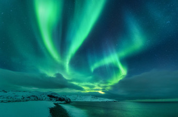 Aurora borealis over ocean. Northern lights in Teriberka, Russia. Starry sky with polar lights and clouds. Night winter landscape with aurora, sea with stones in blurred water, snowy mountains. Travel