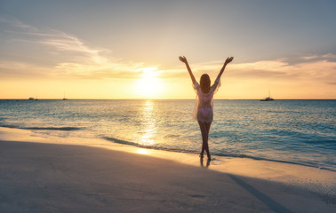 Canvas Print - Beautiful young woman standing in sea with waves on sandy beach against orange sky at sunset. Summer travel. Happy slim girl in white lace dress with raised up arms on the seashore in Zanzibar, Africa