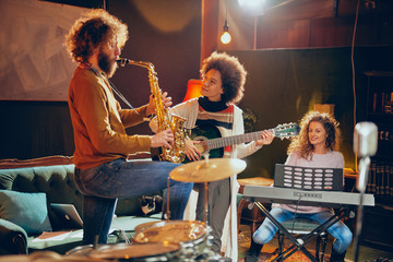 Poster - Mixed race woman playing acoustic guitar while man playing saxophone. Home studio interior. In background woman playing clavier.