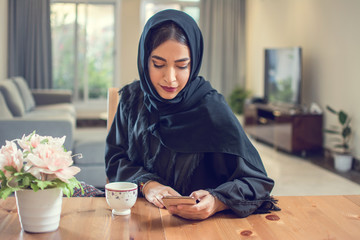 Wall Mural - Beautiful young muslim woman in hijab using smart phone during morning coffee break at home