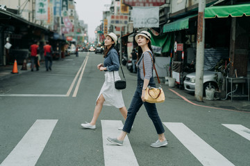 Wall Mural - two happy female friends travelers with bags crossing street together outdoor sunny day in china town. japanese lady travel in chinese city walking on zebra cross in urban. girl wear hats look sky.