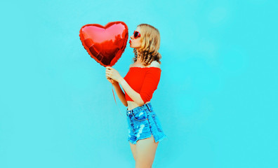 beautiful young woman sending sweet air kiss with red heart shaped balloon in profile on colorful blue background