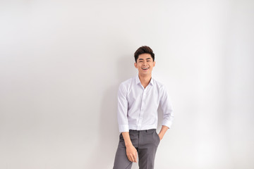 Portrait of happy young man standing against white background. Asian man with hands in pocket leaning against white wall