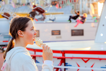 Portrait of cute funny tween girl eating soft vanilla-chocolate ice cream in waffle cone looking at merry-go-round at amusement park. Happy childhood