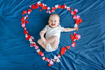 Cute smiling white Caucasian baby girl boy infant with blue eyes four months old lying on bed among many foam paper red pink colorful hearts. View from top above. Happy Valentine day