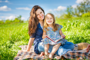 Mother and daughter enjoy reading book sitting on green meadow in the summer park