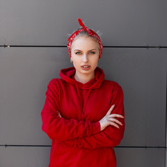 Wall Mural - Stylish pretty young woman in modern fashionable clothes with a red bandanna is standing near a gray modern building on a warm summer day. Beautiful girl model relaxes in the city.