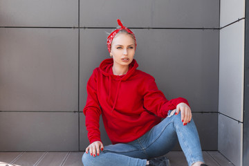 Wall Mural - Attractive modern young woman model in a red stylish bandana in vintage jeans in a fashionable red sweatshirt is resting sitting on the ground near a gray building on a spring day. Cute girl relaxes.