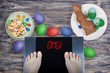 Female feet on digital scales with sign omg! surrounded by Easter food (Easter cake, chocolate Easter bunny, painted eggs). Consequences of overeating and unhealthy lifestile during holidays.
