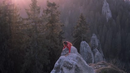 Wall Mural - Young woman hiker with small backpack standing on edge of cliff against a forest background at sunrise