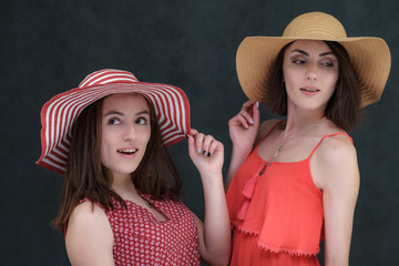 Concept portrait of two stylish brunette sisters on a white and gray background with emotions with hats in summer clothes. Fashionable photo of two beautiful girls