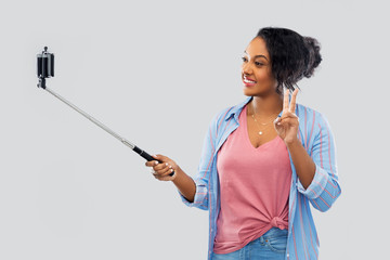 Wall Mural - technology and people concept - happy african american woman taking picture by smartphone on selfie stick and showing peace hand sign over grey background