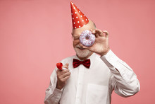 Fun, Joy, Celebration And Holiday Concept. Joyful Positive Mature Retired Male Wearing Red Cone Hat, Bow Tie, White Shirt And Spectacles Relaxing At Birthday Party, Holding Doughnut And Whistle