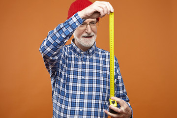 Wall Mural - Isolated image of skilled elderly bearded repairman in glasses, plaid shirt and red hat holding ruler going to take measurements while doing repair works in living room. Architecture and engineering