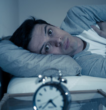 Portrait Of Young Man In Bed Staring At Alarm Clock Trying To Sleep Feeling Stressed And Sleepless