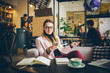 theme modern profession female blogger. Caucasian woman with glasses and jacket sitting inside coffee shop behind wooden table with notebook, laptop and cup coffee. Girl emotion dream, think writer