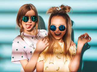 Two young beautiful smiling blond hipster girls in trendy summer colorful T-shirt clothes. Sexy carefree women posing near blue wall in round sunglasses. Positive models having fun and showing tongue