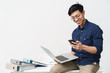 Photo of smiling asian man 20s wearing eyeglasses holding smartphone and laptop while working in office
