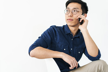 Sticker - Photo of successful asian businessman 20s wearing eyeglasses sitting in chair and talking on smartphone while working in office